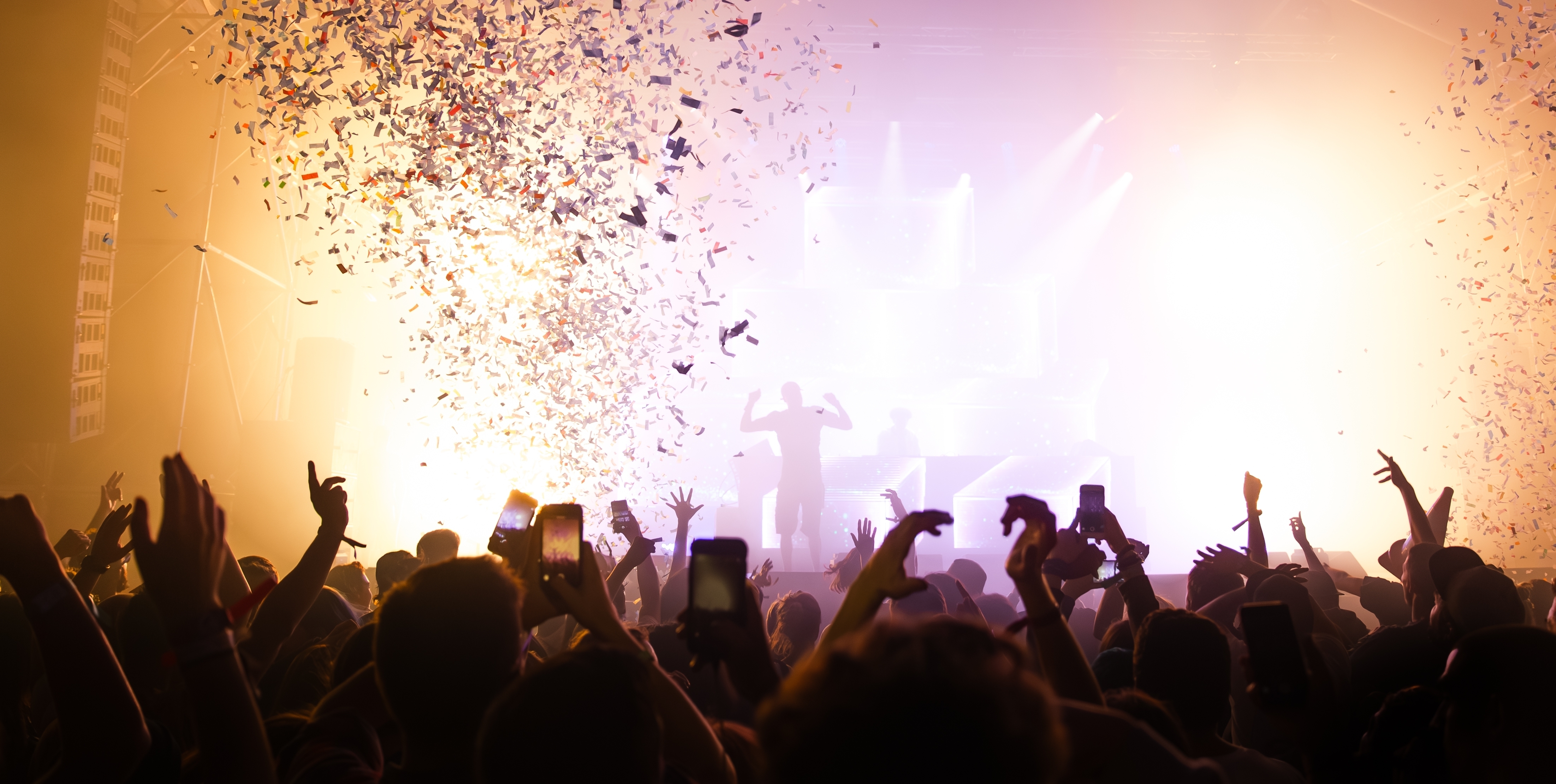 A concert crowd with raised hands beneath pink and purple stage lights